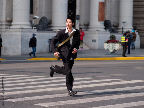 Portrait of handsome Chinese young man with black short hair wearing black blazer running across street with modern city building background in sunny winter day, male fashion, cool Asian young man.