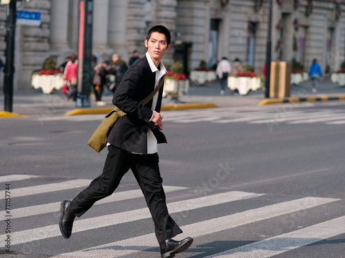 Portrait of handsome Chinese young man with black short hair wearing black blazer running across street with modern city building background in sunny winter day, male fashion, cool Asian young man.