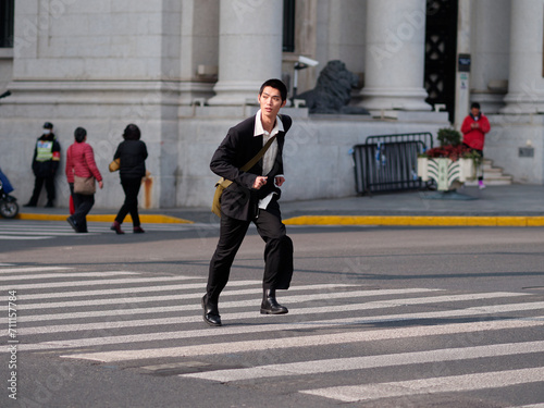 Portrait of handsome Chinese young man with black short hair wearing black blazer running across street with modern city building background in sunny winter day, male fashion, cool Asian young man.