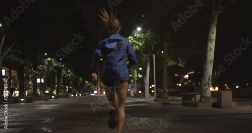 A woman wearing rain rustler running on a street at night. Raining. Back view recorded at 120 fps on cinema camera. photo