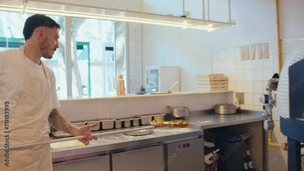 Male chef in apron putting hot pizza on metal plate after baking, using peel to take it off wood-fired oven at work in pizzeria kitchen Male chef in apron putting hot pizza on metal plate after baking, using peel to take it off wood-fired oven at work in pizzeria kitchen