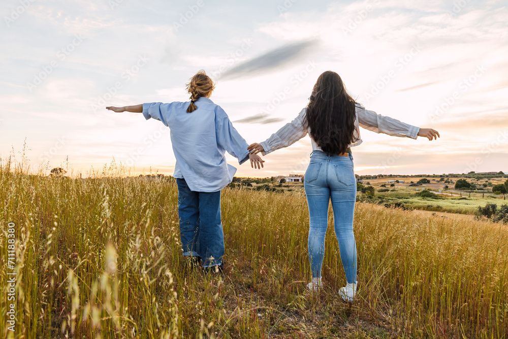 Two young women enjoying nature with the arms open, in a field at sunset