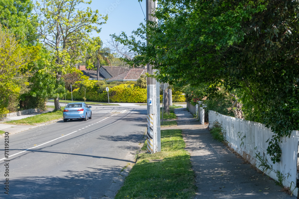 A suburban road with a car, trees, electricity poles, pedestrian sidewalk and houses in the distance in an Australian neighbourhood street. Background texture of a quiet street in residential area.