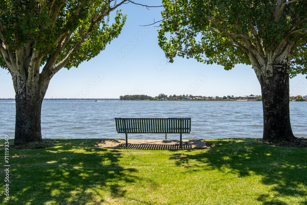 A public seating bench on the lakeside of Lake Mulwala. It is a popular attraction for water sports activities. Background texture of chair by the water. Relaxing vacation vibe. Yarrawonga, Australia.