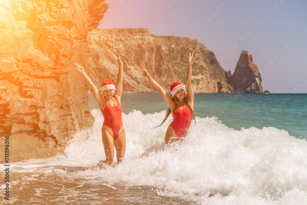 Fototapeta premium Women Santa hats ocean play. Seaside, beach daytime, enjoying beach fun. Two women in red swimsuits and Santa hats are enjoying themselves in the ocean waves and raising their hands up.