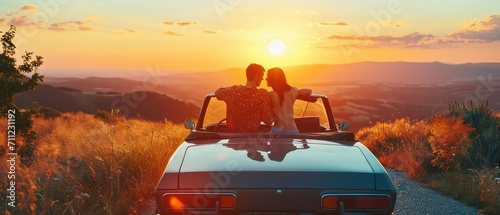 Image of a young couple on a summer road trip, pausing to enjoy a sunset from the hood of their car photo