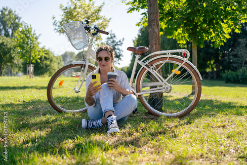 Smiling young hipster woman with bicycle using mobile phone in city park. Talking by video call, using internet