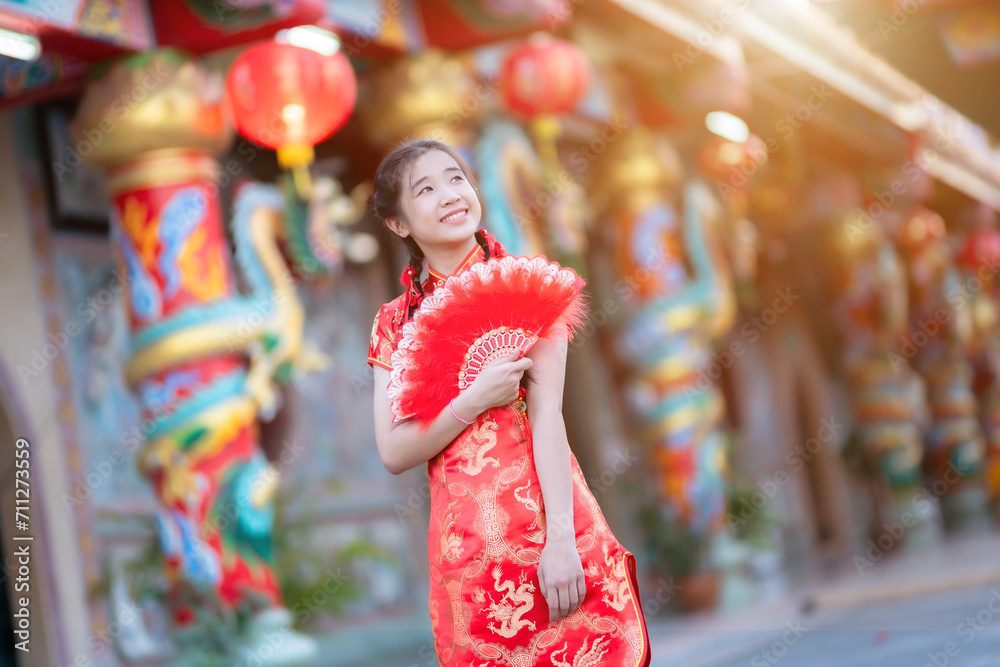 Portrait beautiful cute smiles Asian young woman wearing red traditional Chinese cheongsam decoration and holding a Chinese Fanning for Chinese New Year Festival at Chinese shrine in Thailand