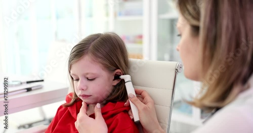 Doctor otorhinolaryngologist examining ear of little girl with otoscope in clinic 4k movie slow motion. Diagnosis and treatment of otitis media and hearing loss in children concept photo