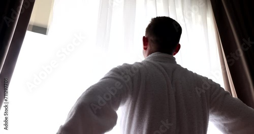 Young man in bathrobe opens curtains at dawn. Rear view of a man with open curtains meets new sunny day in cozy home or hotel