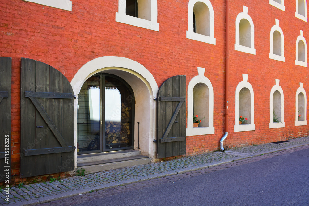 Fototapeta premium Facade of the historic building with small semicircular windows, antique doors with wooden sliding shutters, red brick wall. Ancient architecture, old street. Torun, Poland, August 2023