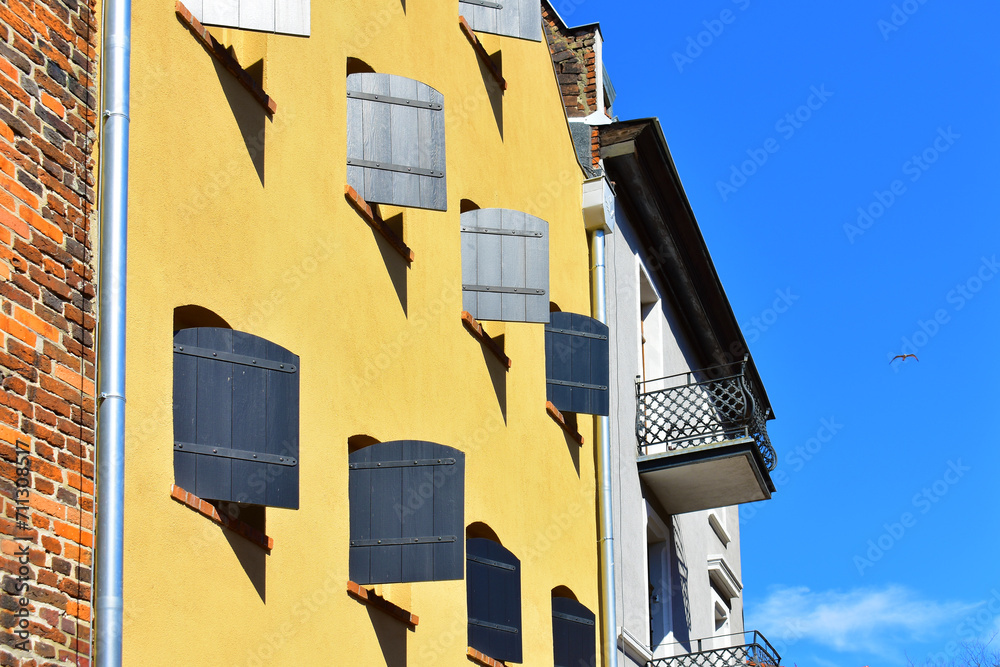 Facade of the historic building with yellow wall and small windows with wooden shutters open. Ancient architecture, rustic style. Torun, Poland, August 2023