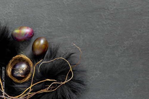 Black fur nest with painted eggs on a dark stone table. Festive Easter banner with branches. Concept design.