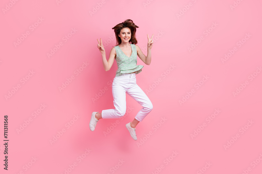 Photo of cheerful good mood girl wear trendy clothes jump up two arms demonstrate v-sign gesture isolated on pink color background