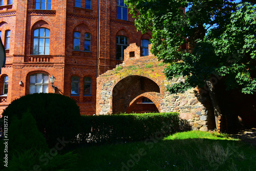 Facade of historic building with red brick walls, tall windows, metal street lamp and large arched passage. Green lawn and tree in the foreground. Ancient architecture. Poland, Torun, August 2023. 