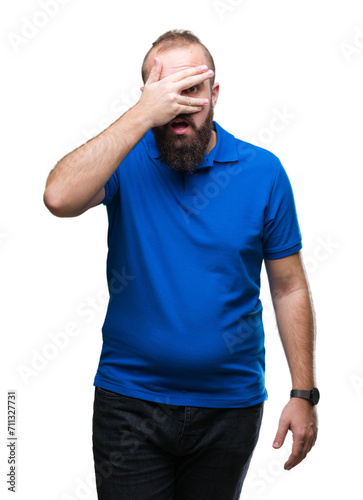 Young caucasian hipster man wearing blue shirt over isolated background peeking in shock covering face and eyes with hand, looking through fingers with embarrassed expression.