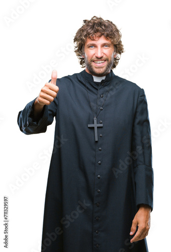 Handsome hispanic catholic priest man over isolated background doing happy thumbs up gesture with hand. Approving expression looking at the camera with showing success.