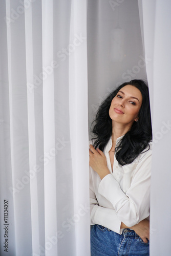In a white shirt and jeans, the lovely young woman poses with poise against delicate white curtains, radiating a captivating aura.