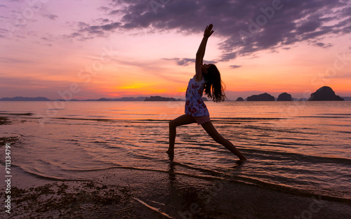 Silhouette of young girl doing Yoga Warrior I Pose on tropical beach with sunset sea sky background. Healthy teenage woman lifestyle balanced practicing meditate and energy yoga. Healthy life Concept.