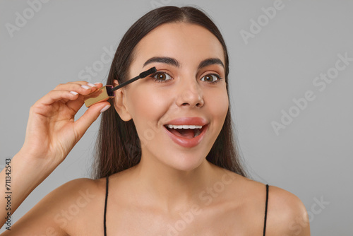 Beautiful young woman applying mascara on grey background