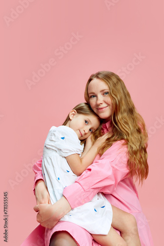 Portrait of happy, beautiful young woman, mother hugging her little daughter against pink studio background. Concept of Mother's Day, International Happiness Day, family, motherhood, childhood
