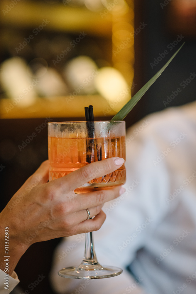 Woman drinking cocktail with slice of orange in a restaurant. Romantic date on Valentine's day. Bride holding fresh juice. Female hand hold cocktail in glass on bar counter at nightclub. Closeup. Woman drinking cocktail with slice of orange in a restaurant. Romantic date on Valentine's day. Bride holding fresh juice. Female hand hold cocktail in glass on bar counter at nightclub. Closeup.