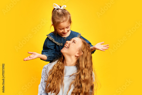 Little girl, daughter surprising her mother against yellow studio background. Celebrating women's holiday. Concept of Mother's Day, International Happiness Day, motherhood, childhood photo