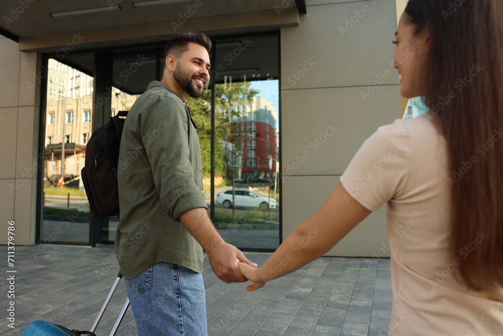 Long-distance relationship. Man with luggage holding hands with his girlfriend near building outdoors Long-distance relationship. Man with luggage holding hands with his girlfriend near building outdoors
