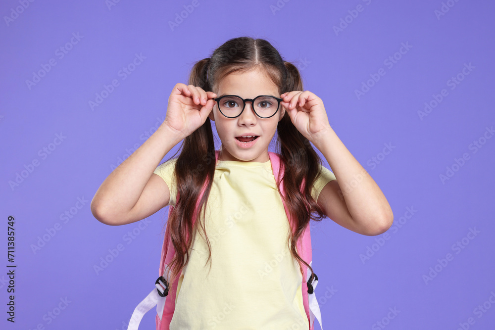 Cute schoolgirl in glasses with backpack on violet background
