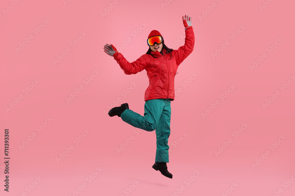 Happy woman in winter sportswear and goggles jumping on pink background