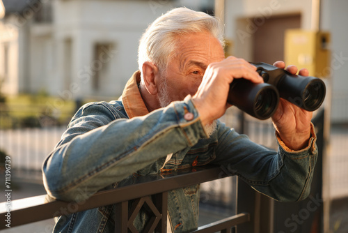 Concept of private life. Curious senior man with binoculars spying on neighbours over fence outdoors photo