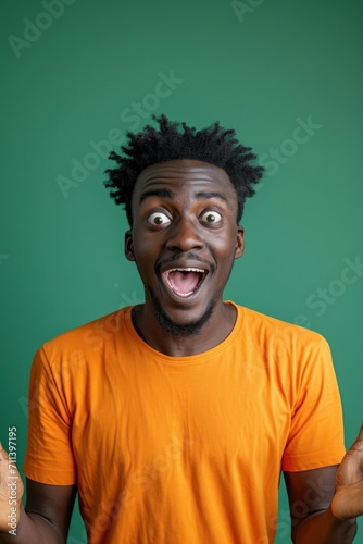 Portrait Young surprised shocked happy man of African American ethnicity. Orange t-shirt green background studio