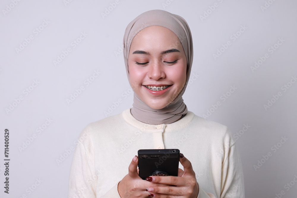 Smiling beautiful Asian woman with braces teeth and hijab using mobile phone received good news isolated over white background Smiling beautiful Asian woman with braces teeth and hijab using mobile phone received good news isolated over white background