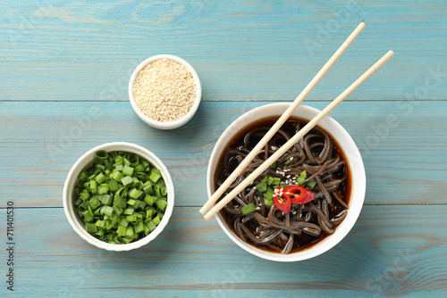 Tasty buckwheat noodle (soba) soup with chili pepper, green onion, sesame and chopsticks on light blue wooden table, flat lay photo
