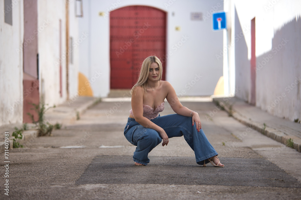 Young, blonde, green-eyed woman with pink top and jeans, posing crouched down on a lonely street in a pretty Andalusian village. Concept of beauty, places, villages, sunny.