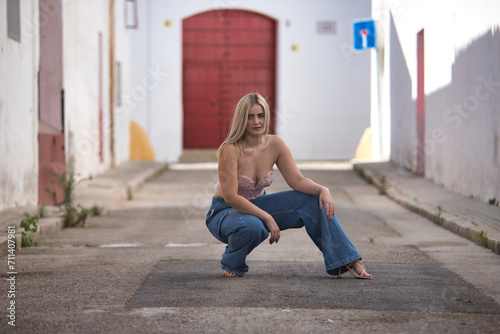 Young, blonde, green-eyed woman with pink top and jeans, posing crouched down on a lonely street in a pretty Andalusian village. Concept of beauty, places, villages, sunny. photo