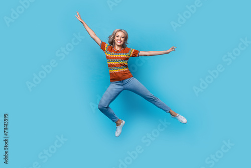 Full length photo of excited cheerful girl dressed print shirt jumping high arms sides isolated blue color background