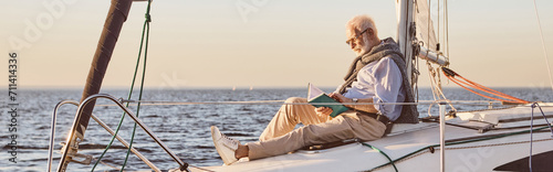 Relaxed senior man reading a book, while sitting on the side of sail boat or yacht deck floating in sea
