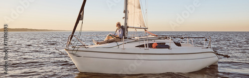 Luxury lifestyle. View of the sail boat or yacht floating in sea with relaxed senior man reading a book, sitting on the deck