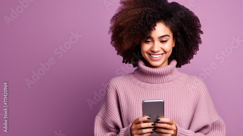 Smiling young multiethnic woman using mobile phone, wearing pink sweater. Portrait cheerful girl with volume curly haired girl on purple background with copy space