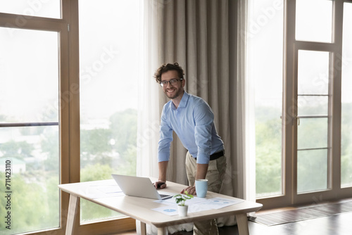 Successful, ambitious young businessman posing for corporate photo standing leaned at workplace desk with laptop and papers. Portrait of small business owner, promoted employee, marketing specialist