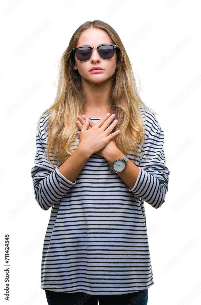 Young beautiful blonde woman wearing sunglasses over isolated background smiling with hands on chest with closed eyes and grateful gesture on face. Health concept. Young beautiful blonde woman wearing sunglasses over isolated background smiling with hands on chest with closed eyes and grateful gesture on face. Health concept.