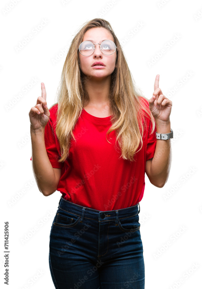 Young beautiful blonde woman wearing glasses over isolated background amazed and surprised looking up and pointing with fingers and raised arms. Young beautiful blonde woman wearing glasses over isolated background amazed and surprised looking up and pointing with fingers and raised arms.