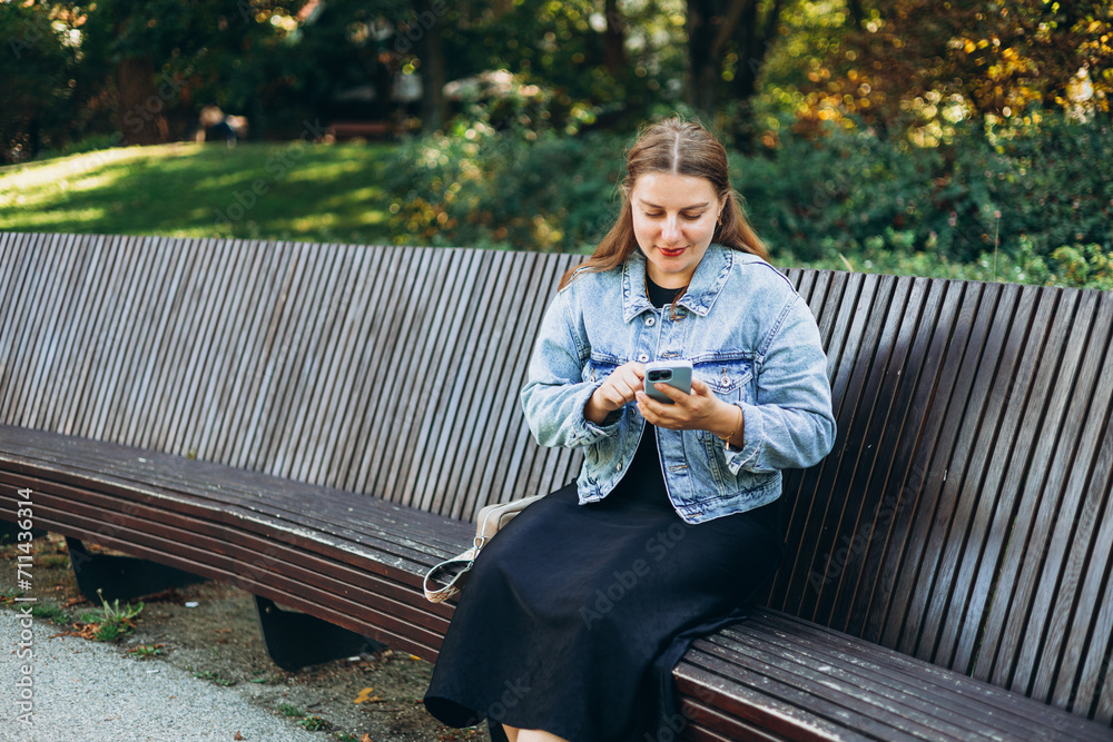 Obraz premium Positive cheerful woman tourist dressed in casual look holding smartphone gadget in hand for communicate on the city street. Happy woman chatting on cell smartphone and relaxing on the bench outdoors