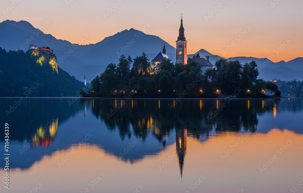 The sun sets on Lake Bled, casting a warm glow on the Pilgrimage Church of the Assumption of Maria and the Bled Castle The sun sets on Lake Bled, casting a warm glow on the Pilgrimage Church of the Assumption of Maria and the Bled Castle