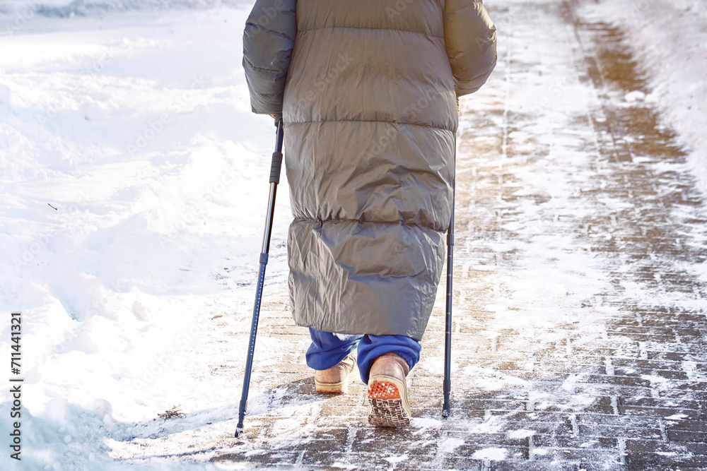 Senior woman practicing nordic pole walking, active lifestyle. Woman trekking with walking sticks in winter day. Alone woman walking along slippery snowy sidewalk with walking cane