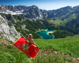 Woman with montenegrin flag at Trnovacko Lake in shape of a heart, surrounded with snowy rocky peaks and green forests. Montenegro, Durmitor National Park, Independence day concept