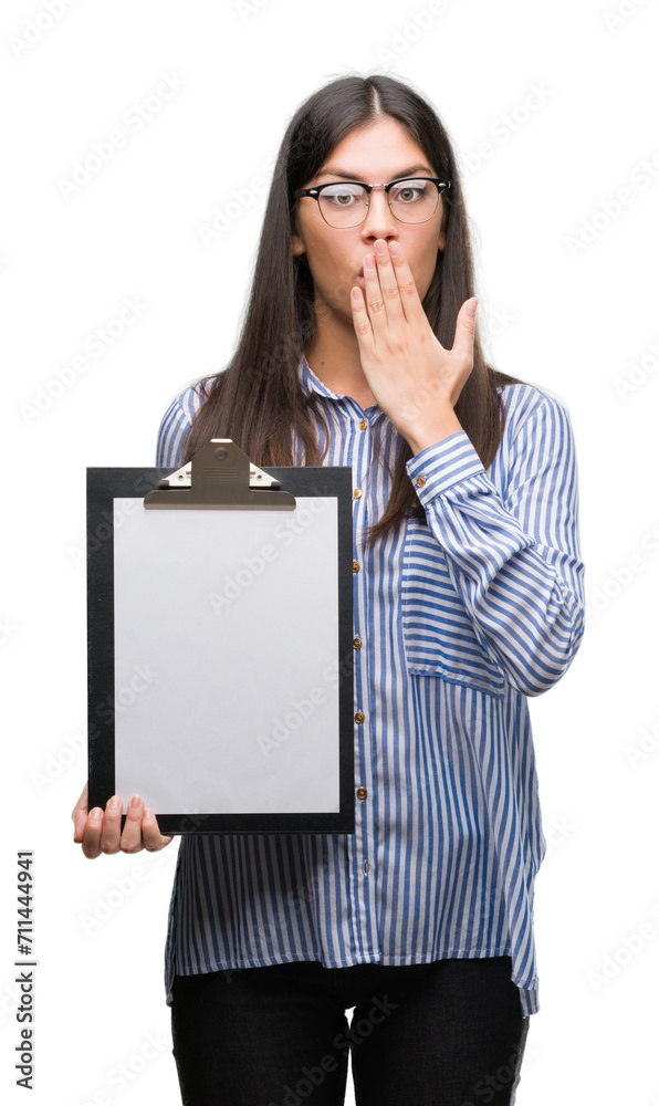 Young hispanic woman holding clipboard cover mouth with hand shocked with shame for mistake, expression of fear, scared in silence, secret concept Young hispanic woman holding clipboard cover mouth with hand shocked with shame for mistake, expression of fear, scared in silence, secret concept