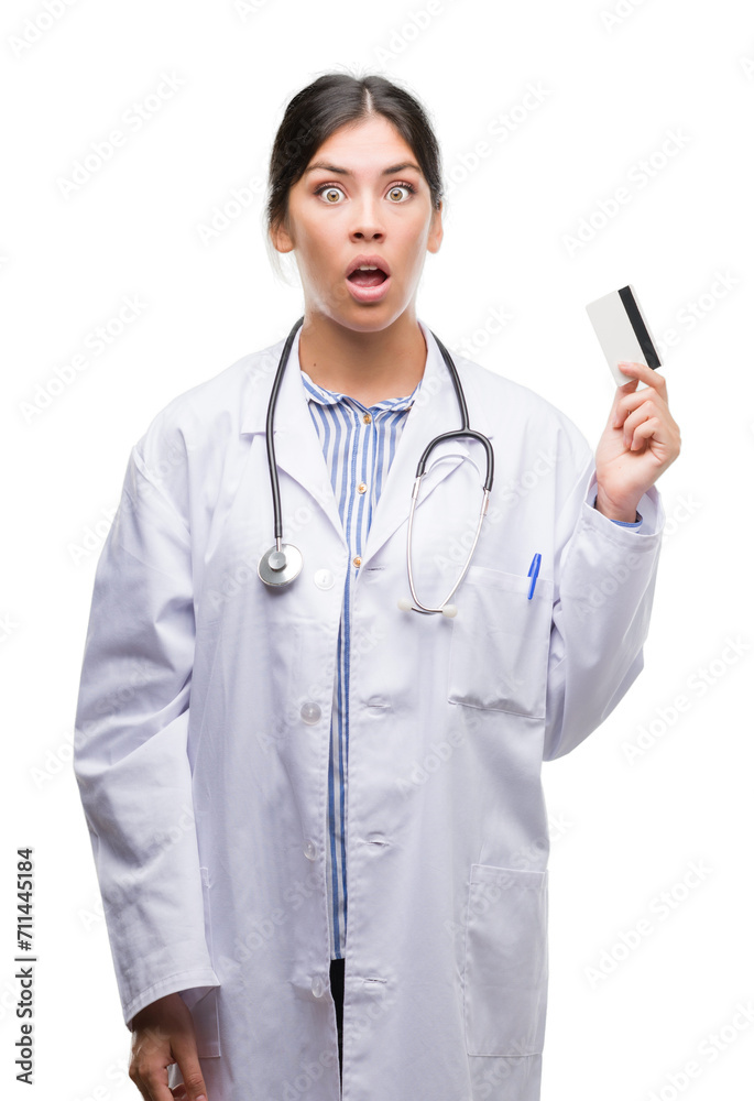 Young hispanic doctor woman holding credit card scared in shock with a surprise face, afraid and excited with fear expression Young hispanic doctor woman holding credit card scared in shock with a surprise face, afraid and excited with fear expression