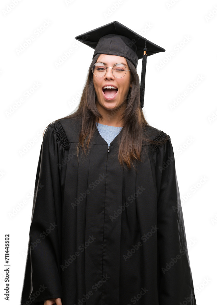 Young hispanic woman wearing graduated cap and uniform winking looking at the camera with sexy expression, cheerful and happy face.